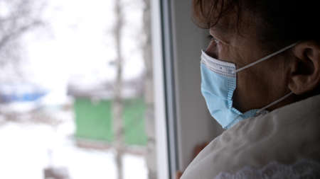 Old despair woman in protective medical mask looking sadly out window, sitting at home in quarantine. Self isolation in pandemic.の写真素材