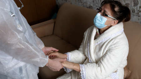 Doctor in a protective suit Helping holding hands Elderly sick woman wearing medical mask At Home. Assistance And Elderly Care Concept. Family Values, Support, Assistance, Seniors Concept.の写真素材