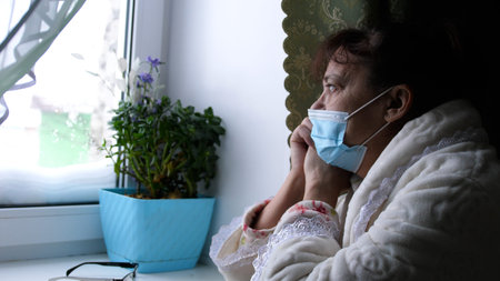 Old despair woman in protective medical mask looking sadly out window, sitting at home in...
