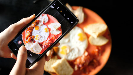 Womens hands take photos of Breakfast food using a smartphone. Fried eggs with fried potatoes, bacon and toastの写真素材