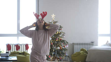 Young beautiful woman wearing reindeer antlers headband and garland dancing next to Christmas treeの写真素材