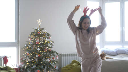 Young beautiful happy woman wearing reindeer antlers headband and galand dancing next to Christmas treeの写真素材