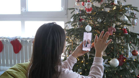 Beautiful Young Smiling Woman near Christmas Tree with Shining Lights at the Background Making Video Message or Selfie Concept of Holidays and New Year in Christmas decoration room.の写真素材