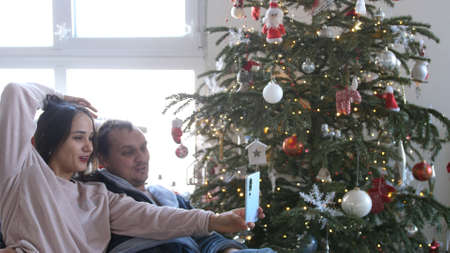Young couple dressed in pajamas having fun and talking with their phone near the Christmas tree. Happy winter holidays and hobby concept.の写真素材