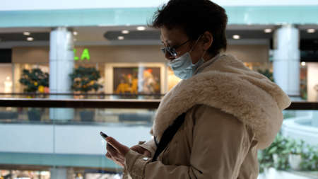 Elderly woman in medical mask and eye glasses uses phone while walking in a shopping mallの写真素材