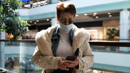 Elderly woman in medical mask and eye glasses uses phone while walking in a shopping mallの写真素材