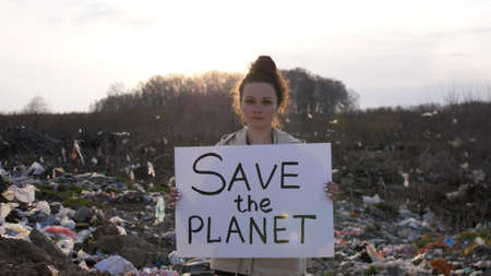 Sad woman volunteer activist stands at big landfill site with garbage with Save the Planet poster demonstrates against protection environment. Pollution and ecology.の写真素材