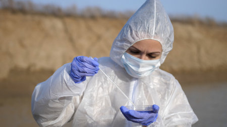 Scientist or ecology expert in protective costume with test tube taking probe for quality control from dirty contaminated water in toxic dangerous zone.の写真素材