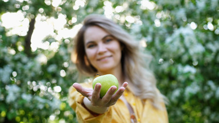 Young pretty woman holding a green apple among blooming apple trees. Organic apple in hand from the tree show to camera. Healthy nutritionの写真素材