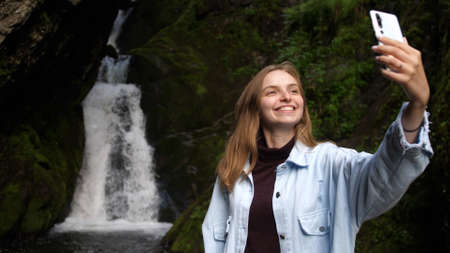 Young pretty woman taking a selfie portrait in front of beautiful waterfall in lush green rainforest, people sharing adventuresの写真素材