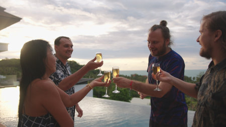 Group of friends having fun at poolside party clinking glasses with white wine by swimming pool on sunny summer day. People toast drinking beverages at luxury villa on tropical vacation.の写真素材