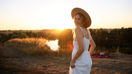Young pretty woman in dress and hat outdoors in field with amazing sunset view enjoying a warm summer day.の写真素材