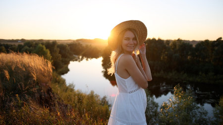 Young pretty woman in dress and hat outdoors in field with amazing sunset view enjoying a warm summer day.の写真素材
