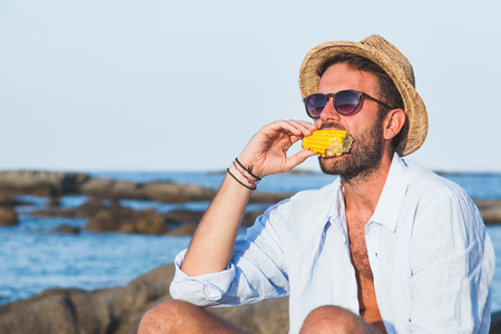 Young man eating corn on the beachの写真素材