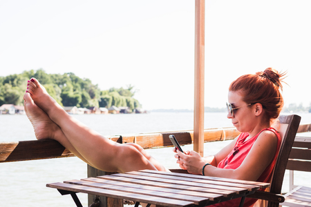 Young woman sitting in a cafe by the river using smartphoneの写真素材