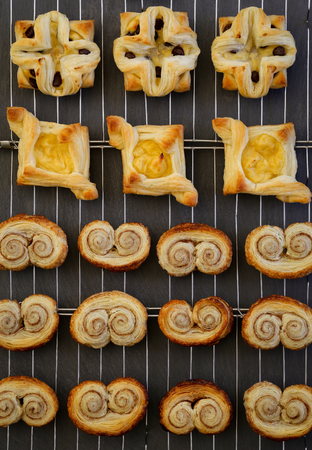 Homemade Gourmet danish puff pastries, flowers with vanilla pastry cream, chocolate chips drops and cinnamon puff pastry scrolls palmiers cooling on wire mesh dark backgroundの写真素材