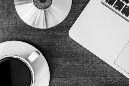 Work in the office, coffee cup and a plate with an optical disk with laptop on the wooden table. In black and white tonesの写真素材