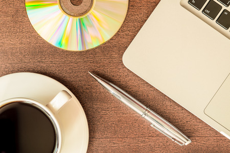 Work in the office, coffee cup and a plate with an optical disk with laptop on the wooden table. In retro tonesの写真素材