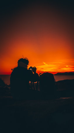Silhouette of a man with a backpack sitting on a rock and looking at the sunsetの写真素材
