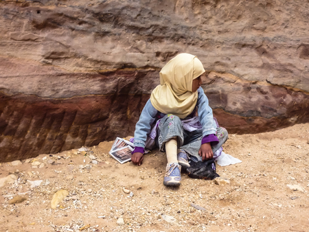 PETRA, JORDAN: A small girl in a national dress of Bedouins sits on the sand on the background of a pink rock, rows lie souvenirs for tourists - postcards with the image of the Mausoleum of El-Khazneのeditorial素材
