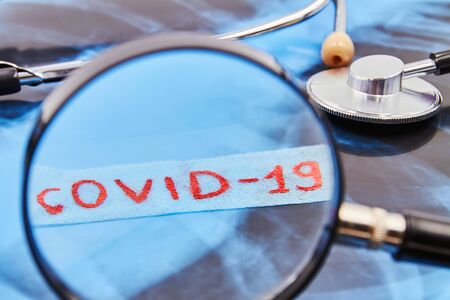Inscription COVID-19 through a magnifying glass on background of stethoscope and lung x-ray. Closeupの写真素材
