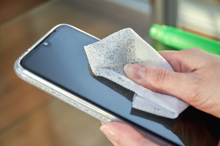 Woman is cleaning a smartphone with a jet of alcohol aerosol hand sanitizer and disposable cloth at home. Precautions regarding virusesの写真素材