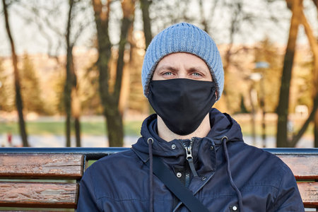 A man in a black protective face mask sitting on a bench in a park. Coronavirus concept. A man wearing protective mask. Closeupの写真素材