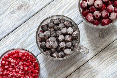 Ripe frozen sweet cherries, red currant and black currant with hoarfrost in the transparent glass cups on blue wooden background. Natural organic healthy food. Top viewの写真素材