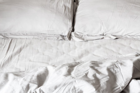 View of an unmade bed with crumpled bed sheet, a blanket and two pillows. Selective focus