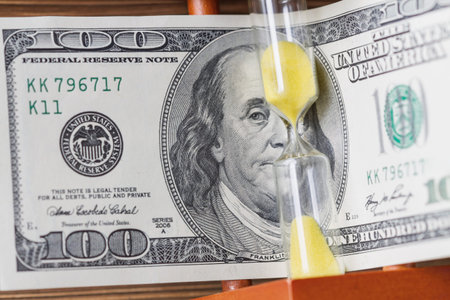 Time is money concept with hourglass and one hundred US dollar bill. Closeup sandglass, selective...