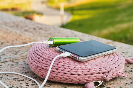 Smartphone and external power bank lie on a womens knitted bag in the park while charging