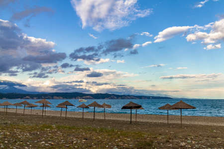 Straw beach umbrellas on the Empty Black Sea coast in Abkhazia, Pitsunda at sunset. Pebble beach, mountains, sea, sky with cloudsの写真素材