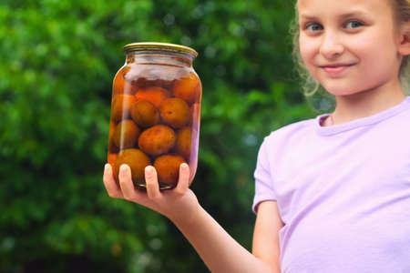 Girls holds a jar with homemade canned plum compote on a background of green foliageの写真素材