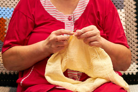 Woman is knitting a beige warm sweater. An aged woman knits on the sofa. A hobby of elderly woman is knitting.の写真素材
