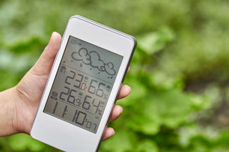 Best personal weather station device with weather conditions inside and outside. A girl holds a gadget in her hand on a background of green foliage.の写真素材