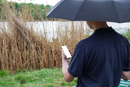 A man under an umbrella uses his smartphone on the shore of a pondの写真素材