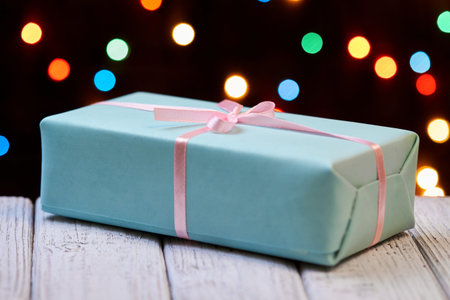 A blue Christmas or New Year gift box with pink ribbon on wooden surface against of colorful lights
