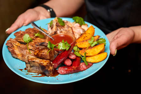 The waiter holds a plate of meat platter with baked potatoes and tomato sauce. Close-up, selective focusの写真素材