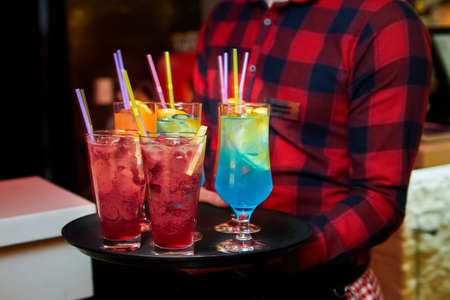 The waiter holds a tray with Multicolored alcoholic and non-alcoholic cocktails with ice and strawsの写真素材