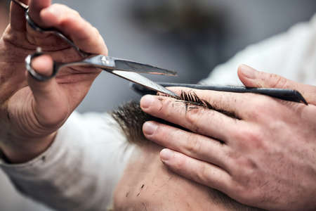 Professional barber making haircut to young man using scissors and comb at barbershop. Close-up, selective focusの写真素材