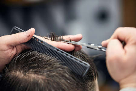 Professional barber making haircut to young man using scissors and comb at barbershop. Close-up, selective focusの写真素材