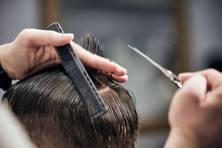 Professional barber making haircut to young man using scissors and comb at barbershop. Close-up, selective focusの写真素材