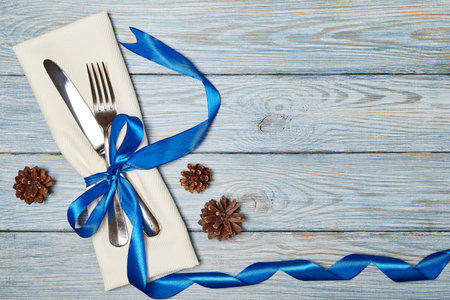 Knife and fork on napkin tied with blue ribbon on wooden background with pine cones