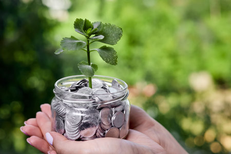 Girl holding in her hands a young plant in a glass jar with coins as a concept for investing and savingの写真素材