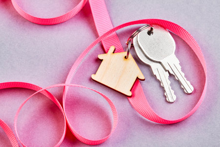 Trinket in the form of a wooden house with the keys to the apartment on purple background with pink ribbonの写真素材