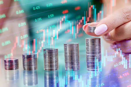 Woman puts coins to a stack of coins and Rising stock market chart as a concept for investing and savingの写真素材