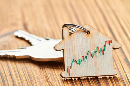 Trinket in the form of a wooden house with the keys to the apartment on wooden background with real estate growth chartの写真素材