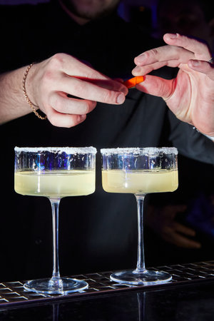 Close-up of bartender sprinkling to glass with cold alcohol cocktail with piece of orange citrus...
