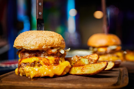 Burger with two beef patties, a wheat bun and cheese, with a knife stuck into it, served with baked potatoesの写真素材