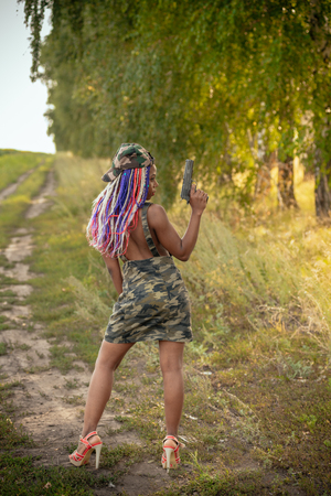 portrait of a beautiful young African American girl with a dreddy hairdress on a park background, nature, deciduous forest, with a gun in her hands. concept of photo walks, female self-defenseの写真素材